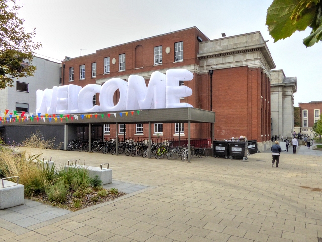 'Welcome' sign displayed outside university, with students and staff walking past