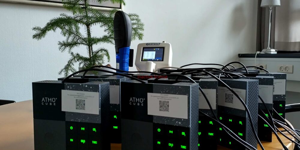 A group of AtmoCube air quality monitoring devices connected with cables on a table, with a GrayWolf device and a small plant in the background.