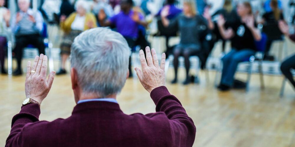 This image depicts an elderly man standing in front of a blurred crowd.