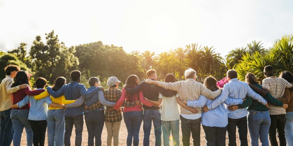 A diverse group of people standing outdoors with their arms around each other, facing the sun, symbolizing unity and togetherness.